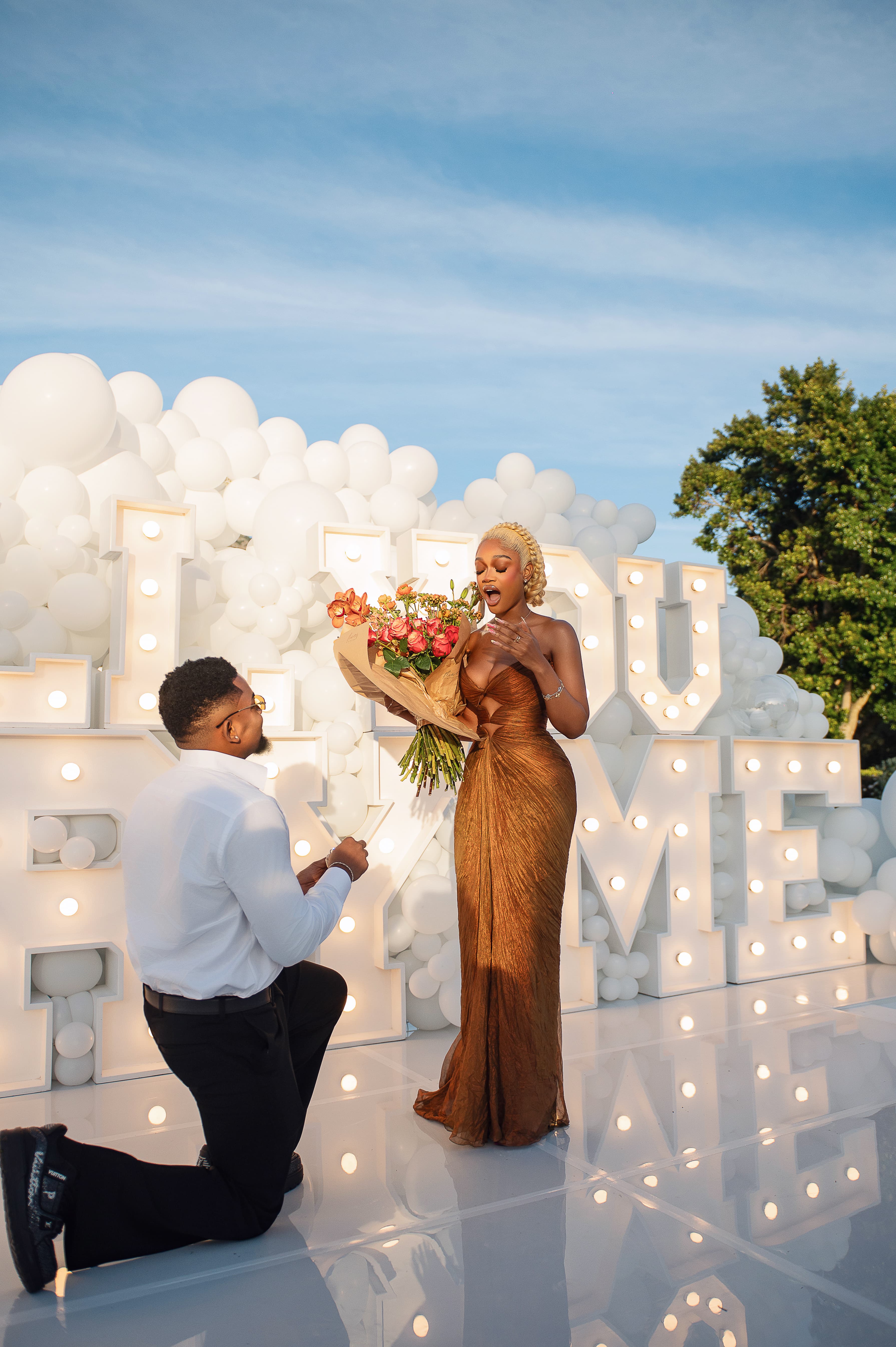 Pascal on one knee with a bouquet; Diana's hand at her mouth, in front of a lit MARRY ME sign